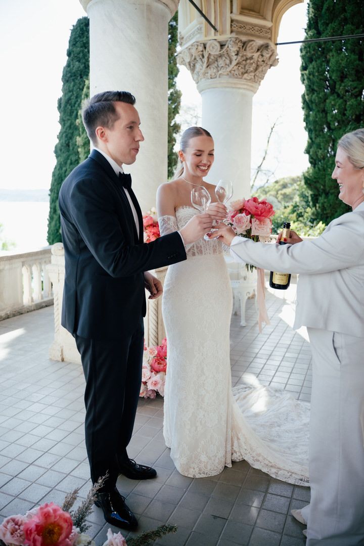 A celebratory toast moment during a wedding on an elegant columned terrace with pink floral arrangements.