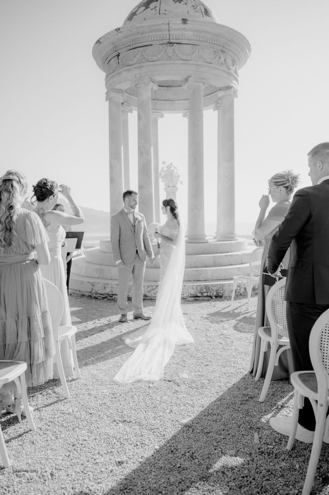 Wedding ceremony takes place under classical white stone gazebo with guests seated on chairs.