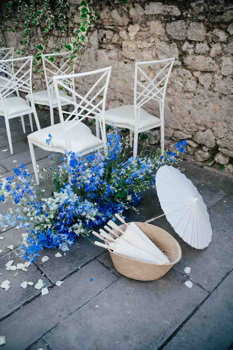 White garden chairs with delicate blue flowers and a white paper parasol arranged against a stone wall.