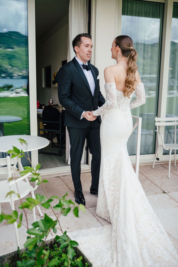 A bride in a fitted lace wedding gown stands with a formally dressed guest on an outdoor terrace with mountain views.