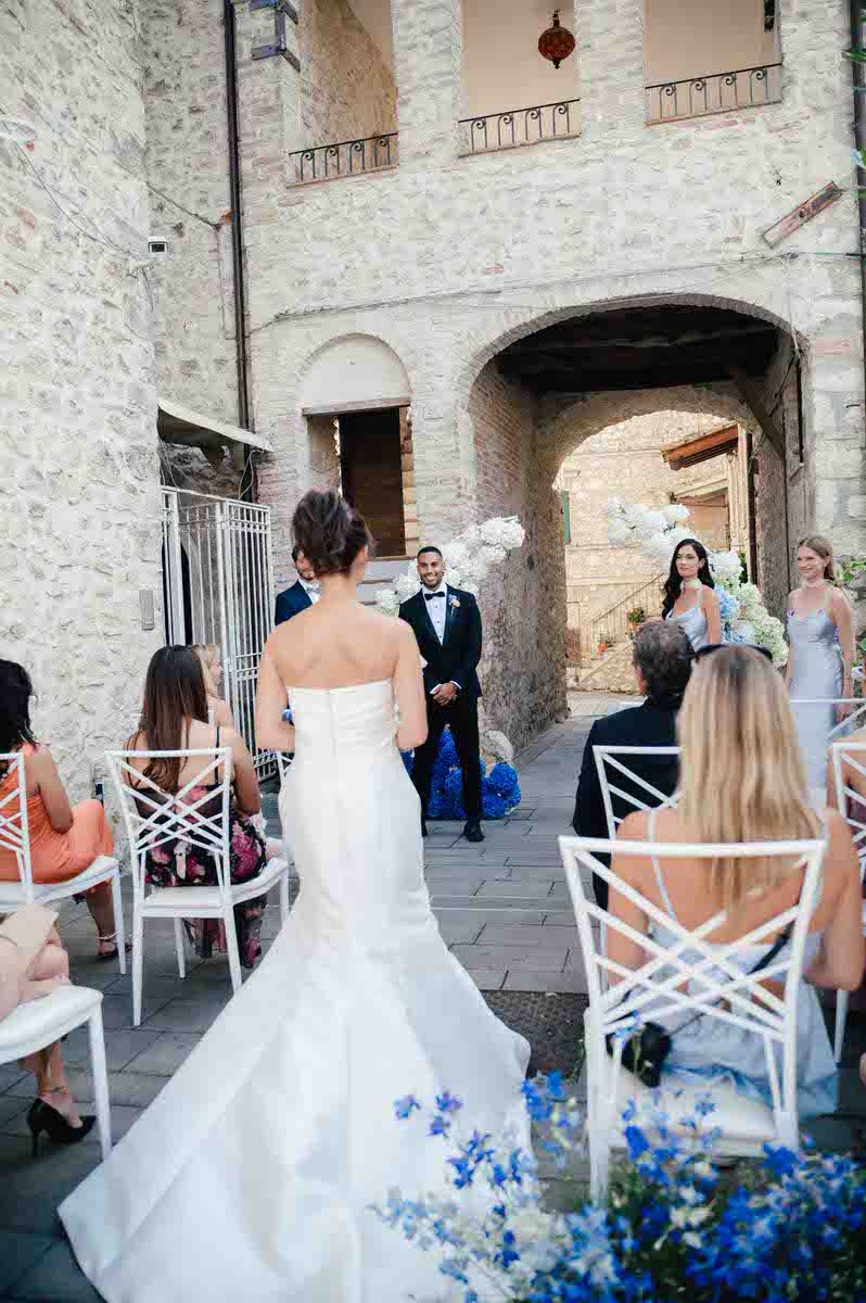 A wedding ceremony in progress at a rustic stone venue decorated with blue flowers and white chairs.