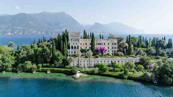 Luxurious Italian villa with cypress trees sits along Lake Garda's shoreline against dramatic mountain backdrop.