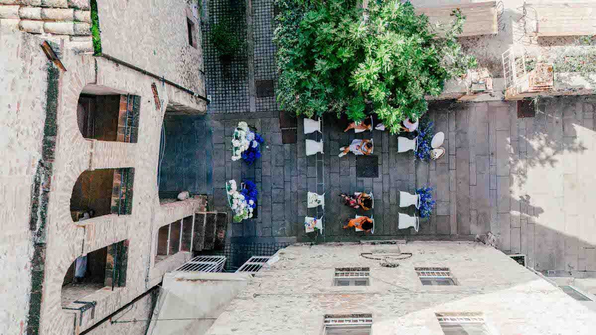 Aerial view of a wedding ceremony setup in a stone courtyard with white chairs and blue decorations.
