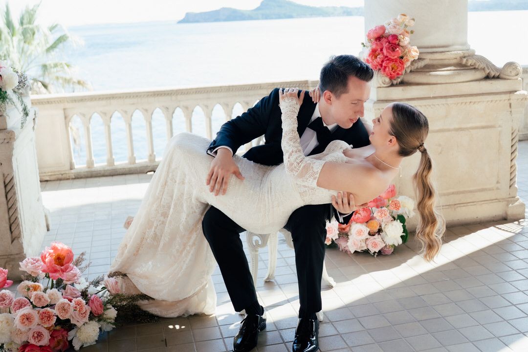 A romantic wedding moment on an elegant balcony overlooking the ocean with pink floral decorations.
