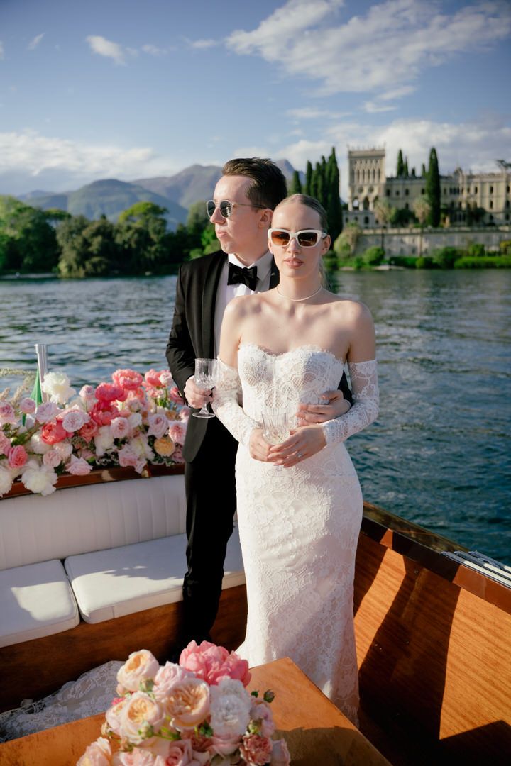 A wedding couple stands on a wooden boat adorned with pink flowers on Lake Como with Villa Balbianello in the background.