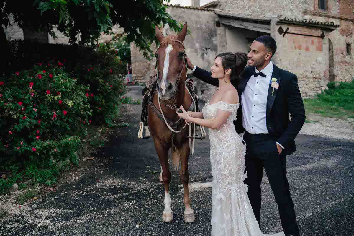 Elegant couple poses with brown horse outside historic stone building with climbing roses.