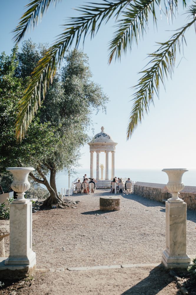 Palm fronds frame classic stone gazebo on coastal cliff with guests gathering for ceremony.