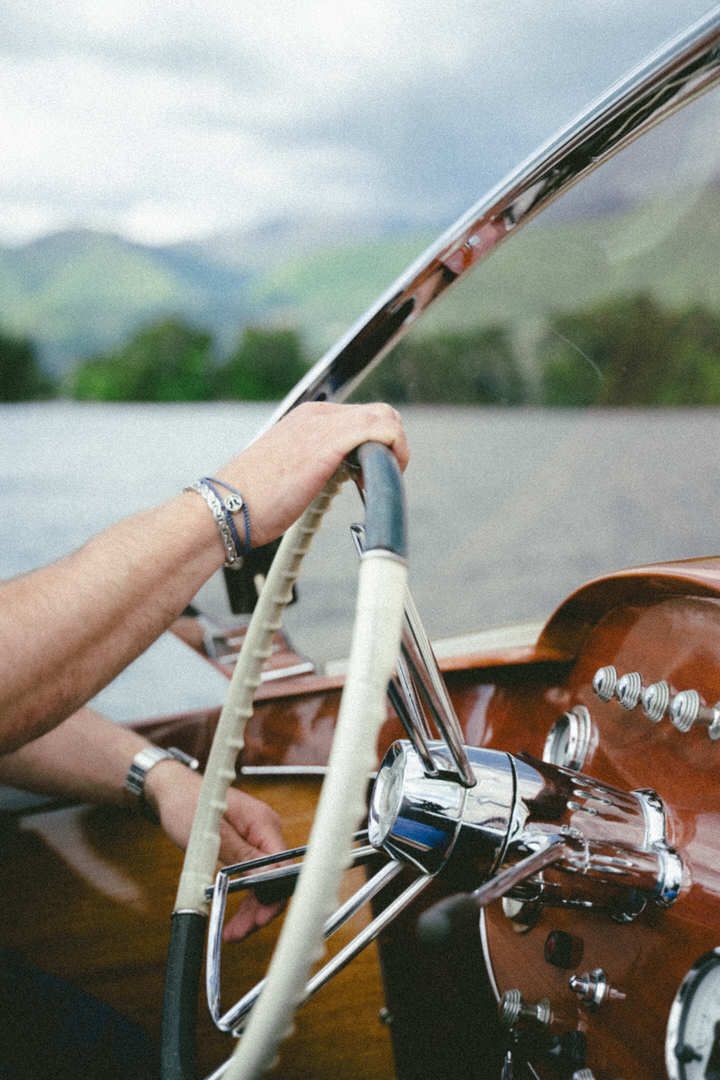 Close-up of hands steering a wooden boat wheel on a lake with mountains in the background.