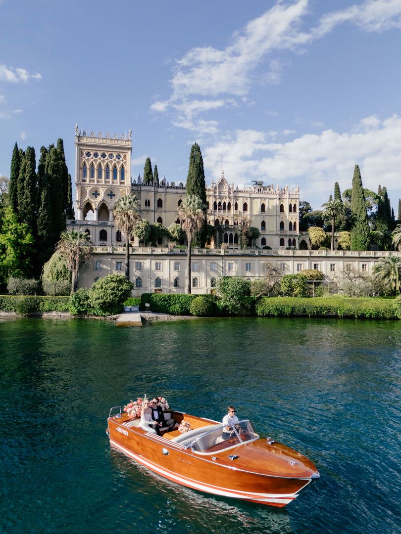Vintage wooden boat cruising past historic Italian villa on Lake Garda with cypress trees along the waterfront.