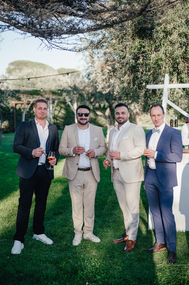 A group of people in light-colored suits gather outdoors holding drinks during what appears to be a social event.