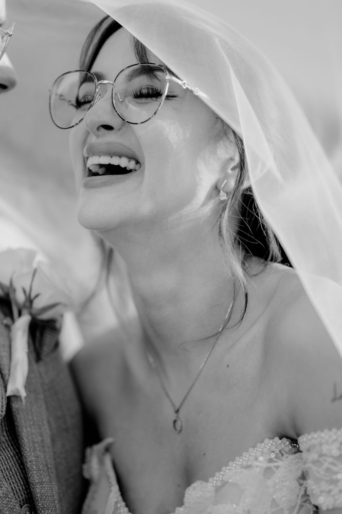 A black and white portrait of a joyful bride wearing glasses and a flowing veil.