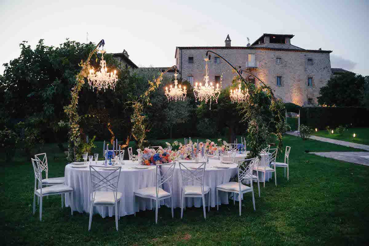 Romantic outdoor wedding reception setup with white tables and chairs under glowing chandeliers.
