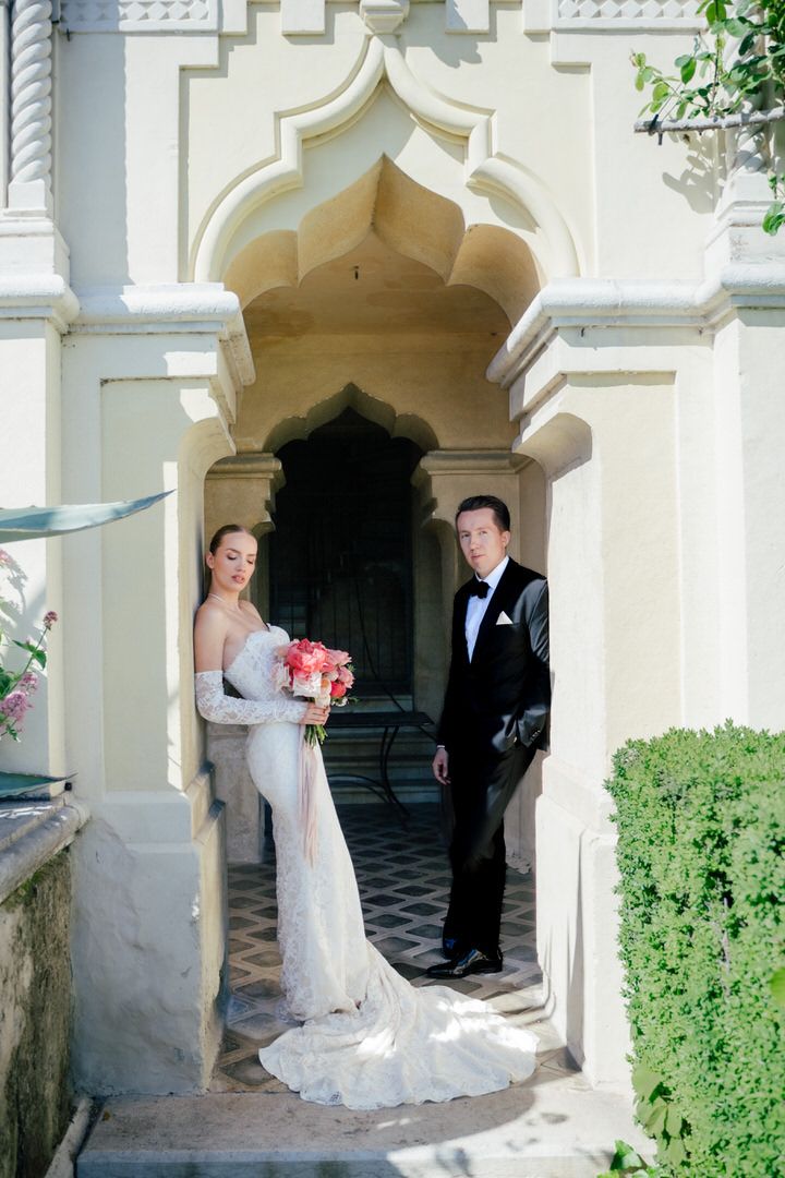 A couple stands in an ornate archway of a white stucco building, with greenery and elegant architectural details visible.