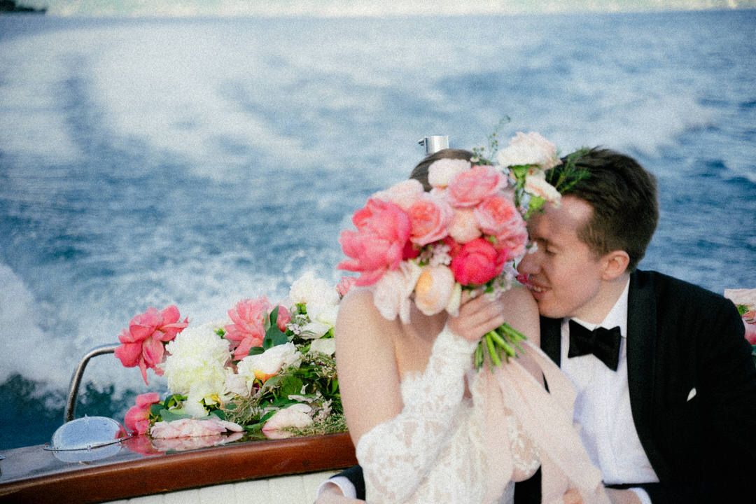 A romantic wedding couple share a kiss aboard a boat with pink and white floral arrangements in the foreground.