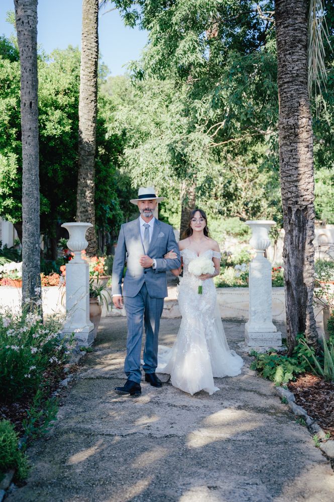 Wedding couple poses on garden path lined with palm trees and white pillars.