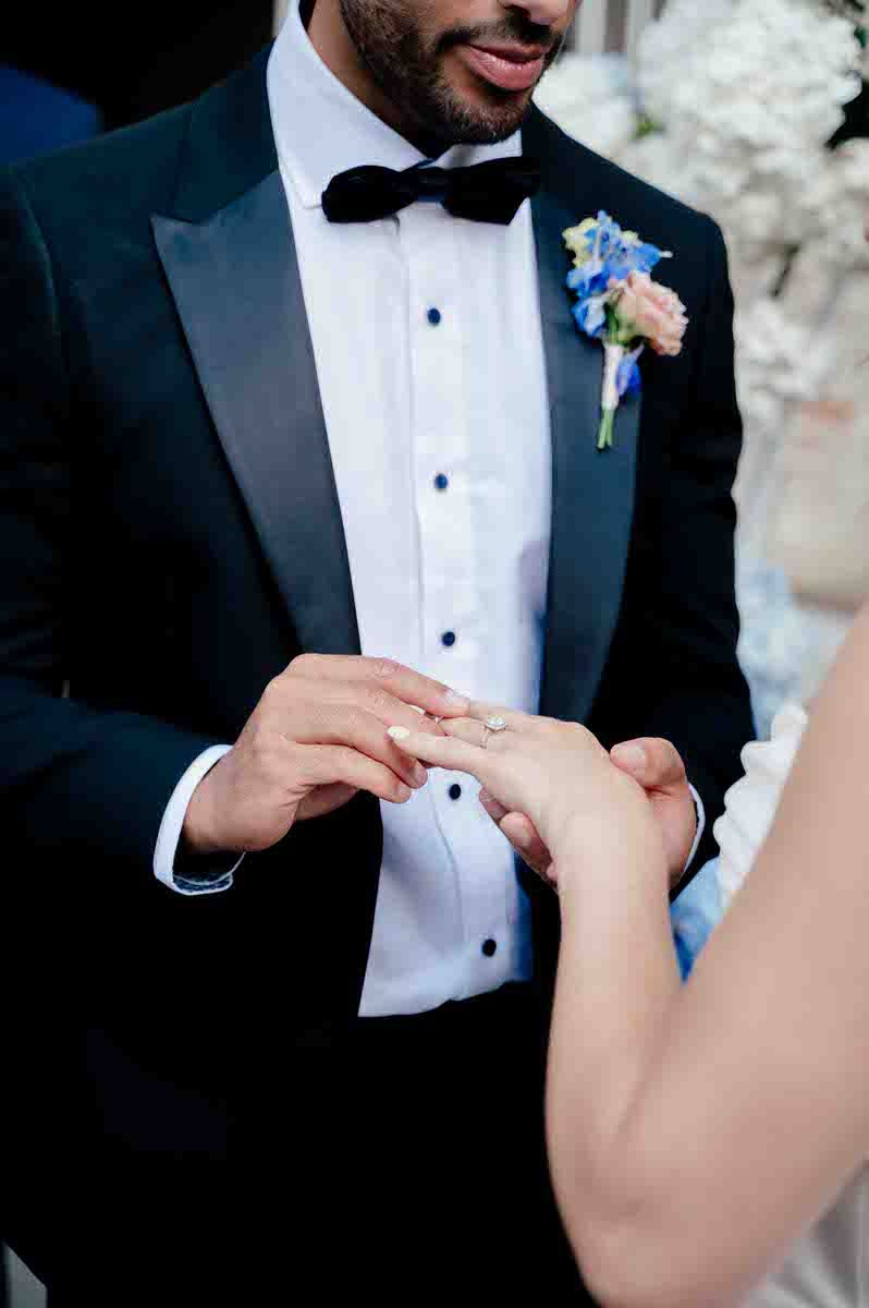 Close-up of hands exchanging rings during wedding ceremony with groom in black tuxedo.