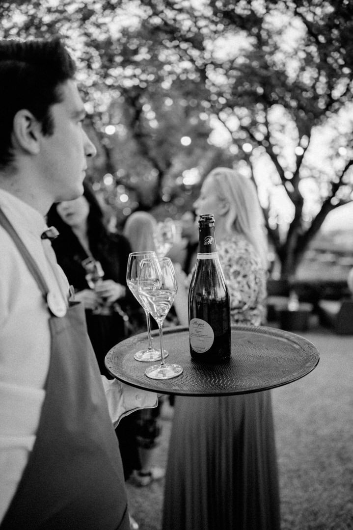 Black and white photo of a busy outdoor party scene with a server holding a tray of wine glasses and bottles.