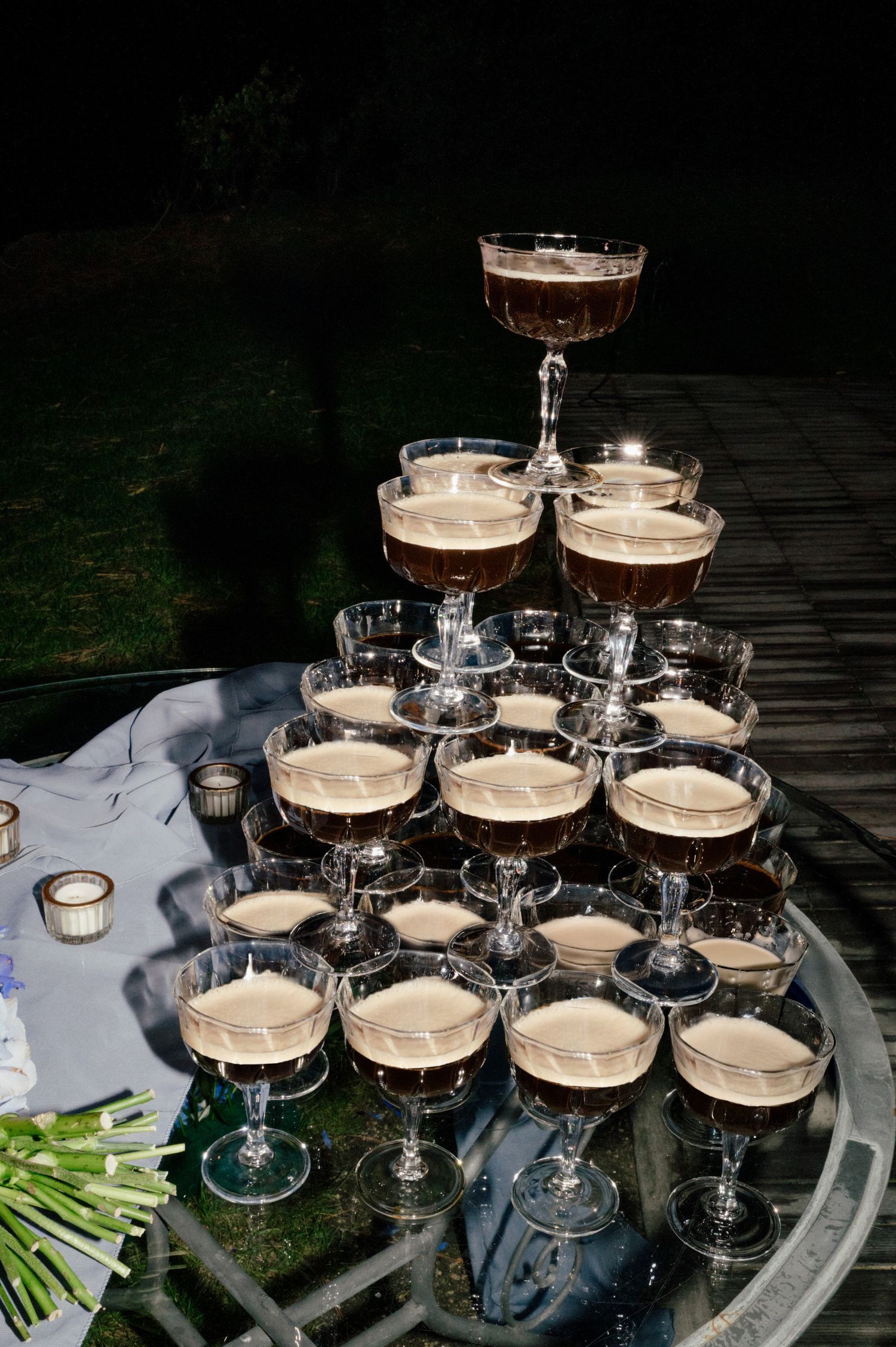 Multiple glass champagne coupes arranged in rows and filled with creamy cocktails on an outdoor table at dusk.