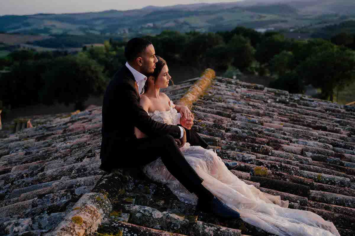 Couple sits romantically on rustic terracotta roof tiles overlooking Italian countryside at sunset.