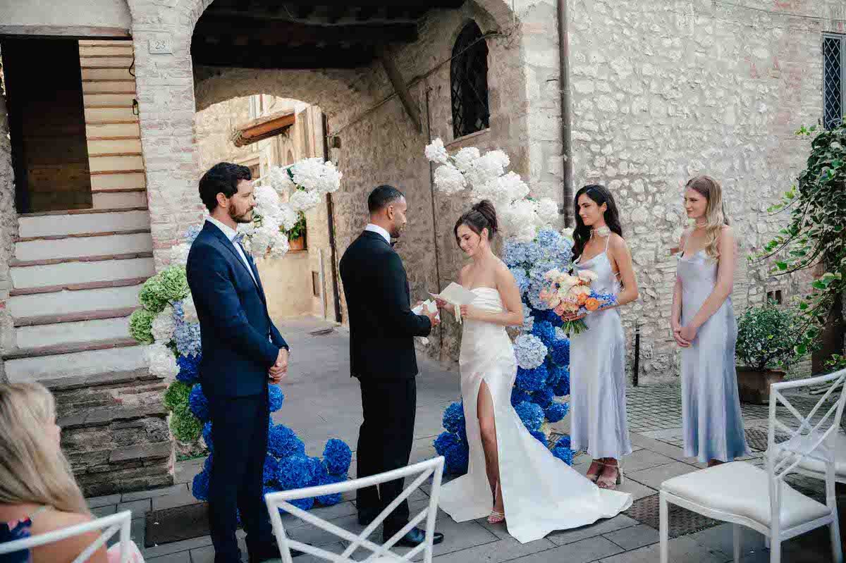 Wedding ceremony taking place in an intimate stone courtyard with white chairs and blue floral accents.