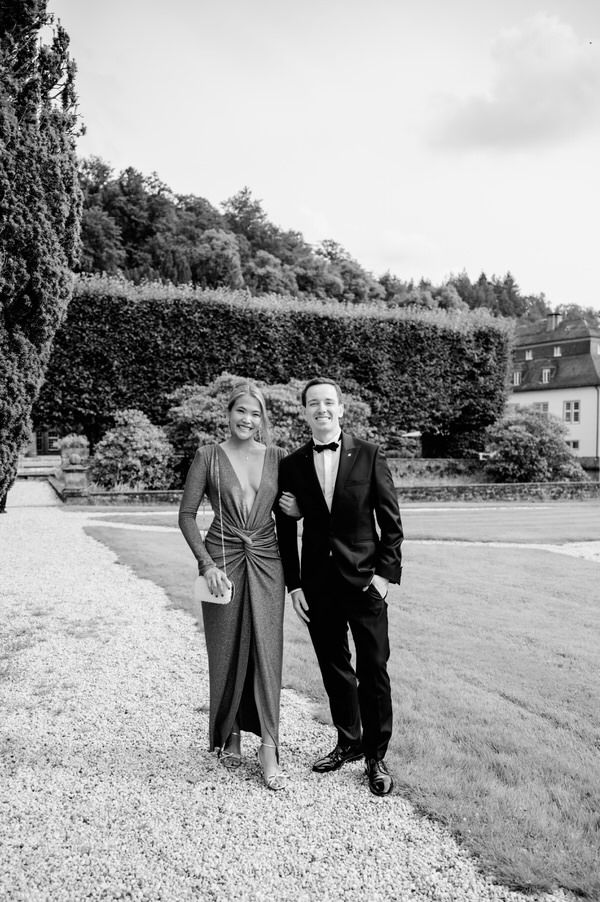 A couple in formal evening attire posing together on a gravel path in a garden setting, captured in black and white.