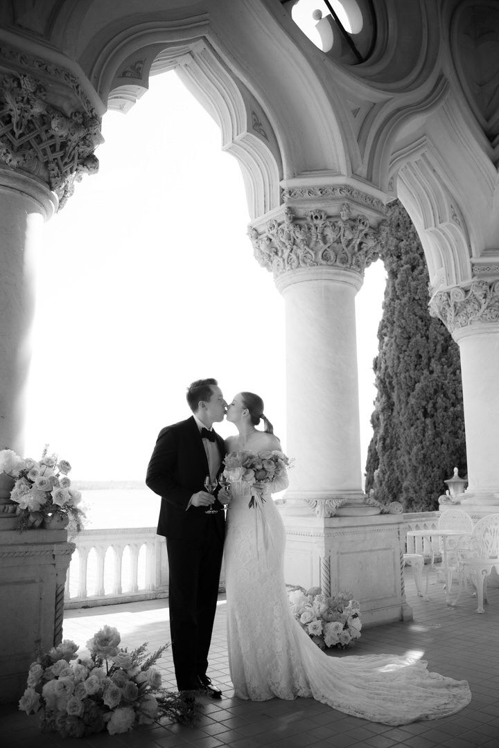 Elegant black and white wedding photo showing couple embracing under ornate classical columns with floral arrangements.