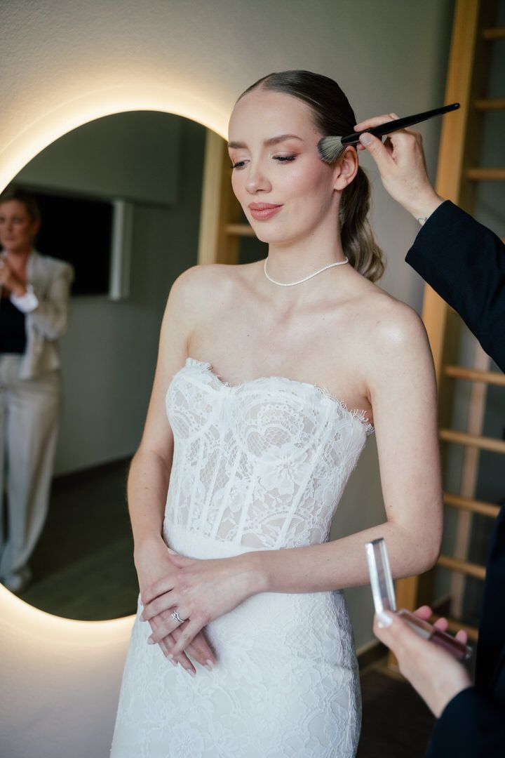 A bride in a white lace strapless wedding gown getting her final touches of makeup done before the ceremony.