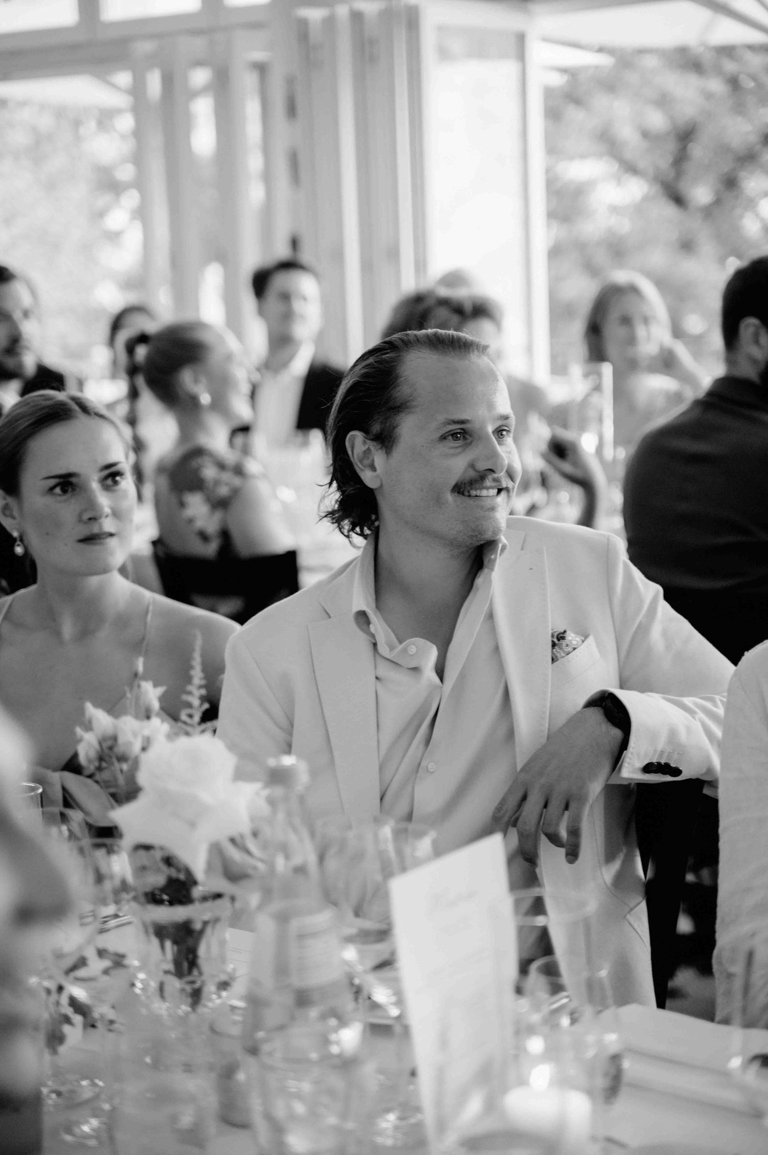 Black and white photo of a wedding reception with guests seated at an elegant table with glassware and floral arrangements.