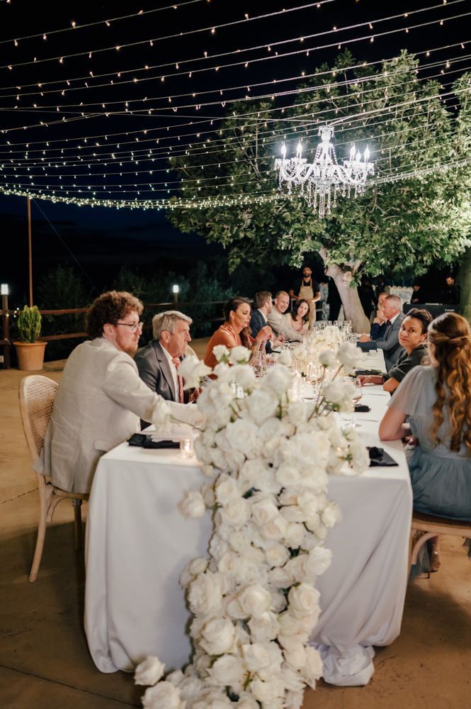 Elegant outdoor wedding reception with string lights overhead and white floral table runner under a crystal chandelier.