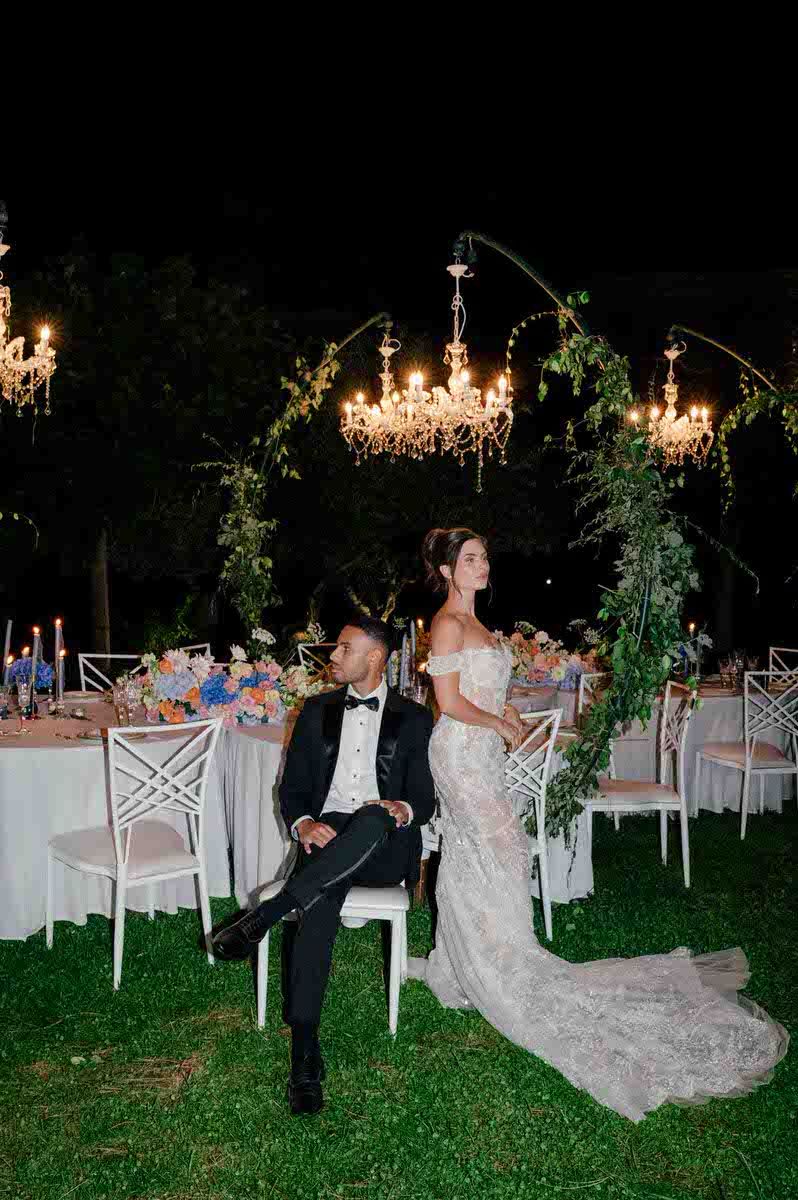 Elegant outdoor wedding reception with chandeliers and greenery at night.