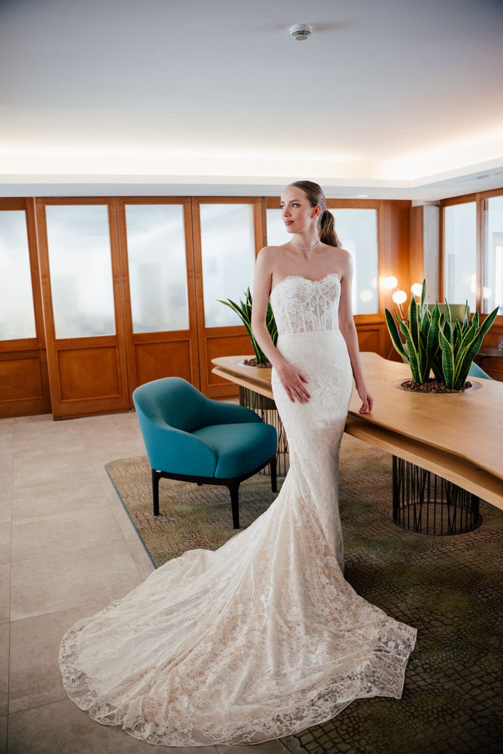 A stunning lace mermaid wedding gown with long train displayed in a modern room with teal accent chair.