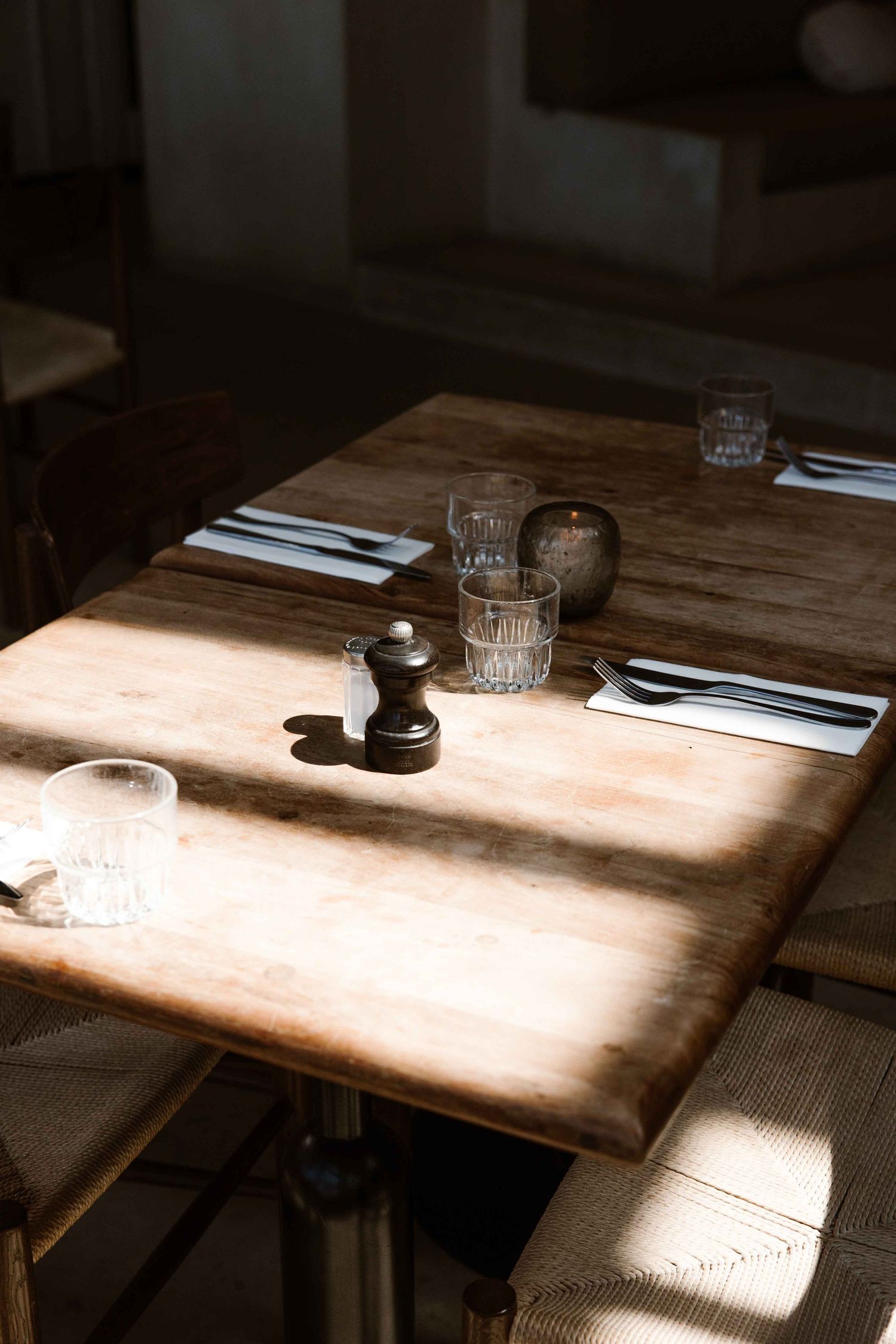 A rustic wooden restaurant table set with glasses and cutlery illuminated by natural sunlight streaming across the surface.