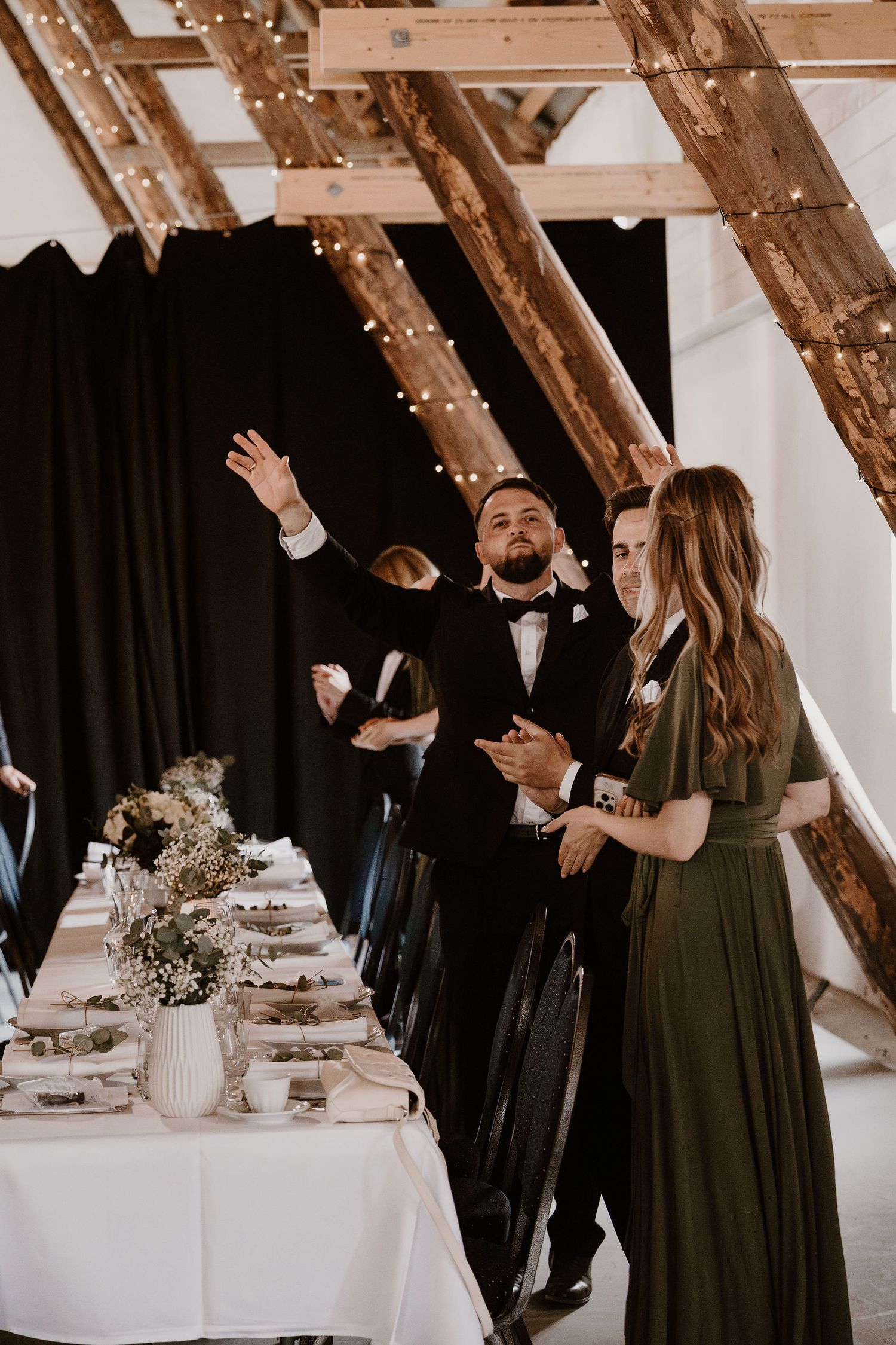 Wedding guests in formal attire raise their hands in celebration at an elegant reception with wooden beams and white decor.