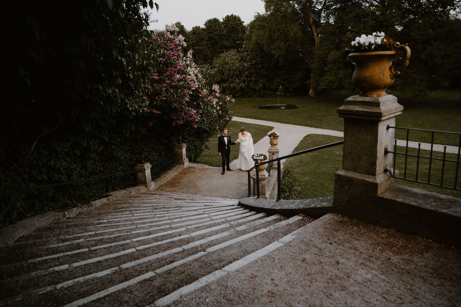 A couple stands together on ornate stone steps in a romantic garden setting at dusk.