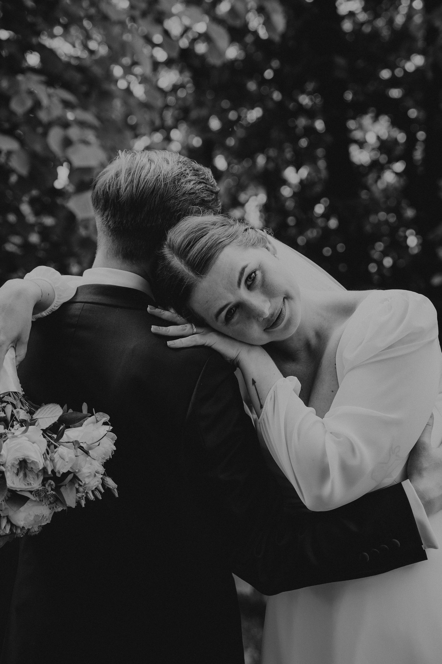 Black and white candid wedding photo showing a bride in white dress embracing groom in suit from behind against bokeh background.
