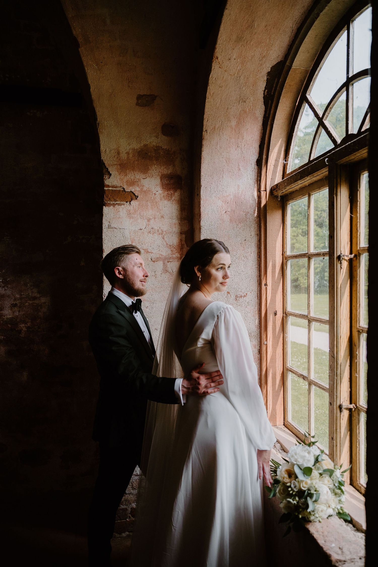 Romantic newlywed couple stands together at rustic window in brick room creating moody wedding portraits.