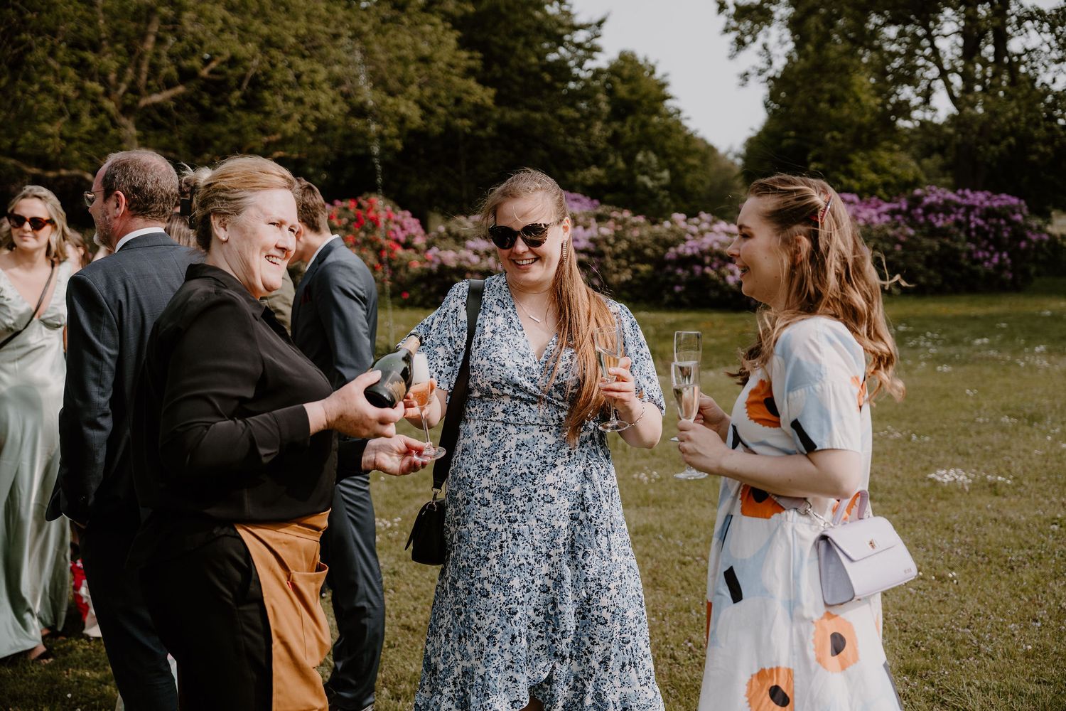 A group of wedding guests enjoying drinks and conversation on a sunny lawn with blooming flowers in the background.