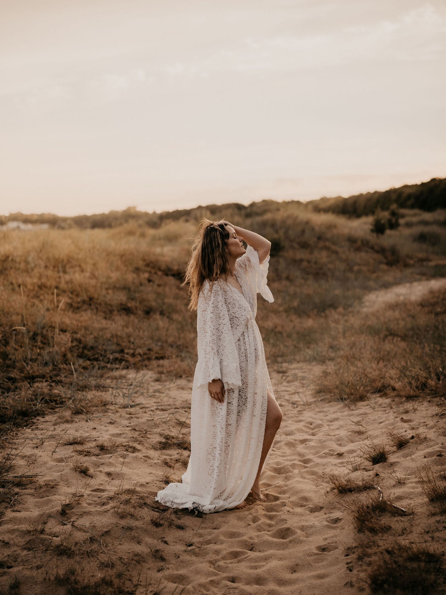 A lone figure in a flowing white lace dress poses on sandy dunes at golden hour in a dreamy outdoor setting.