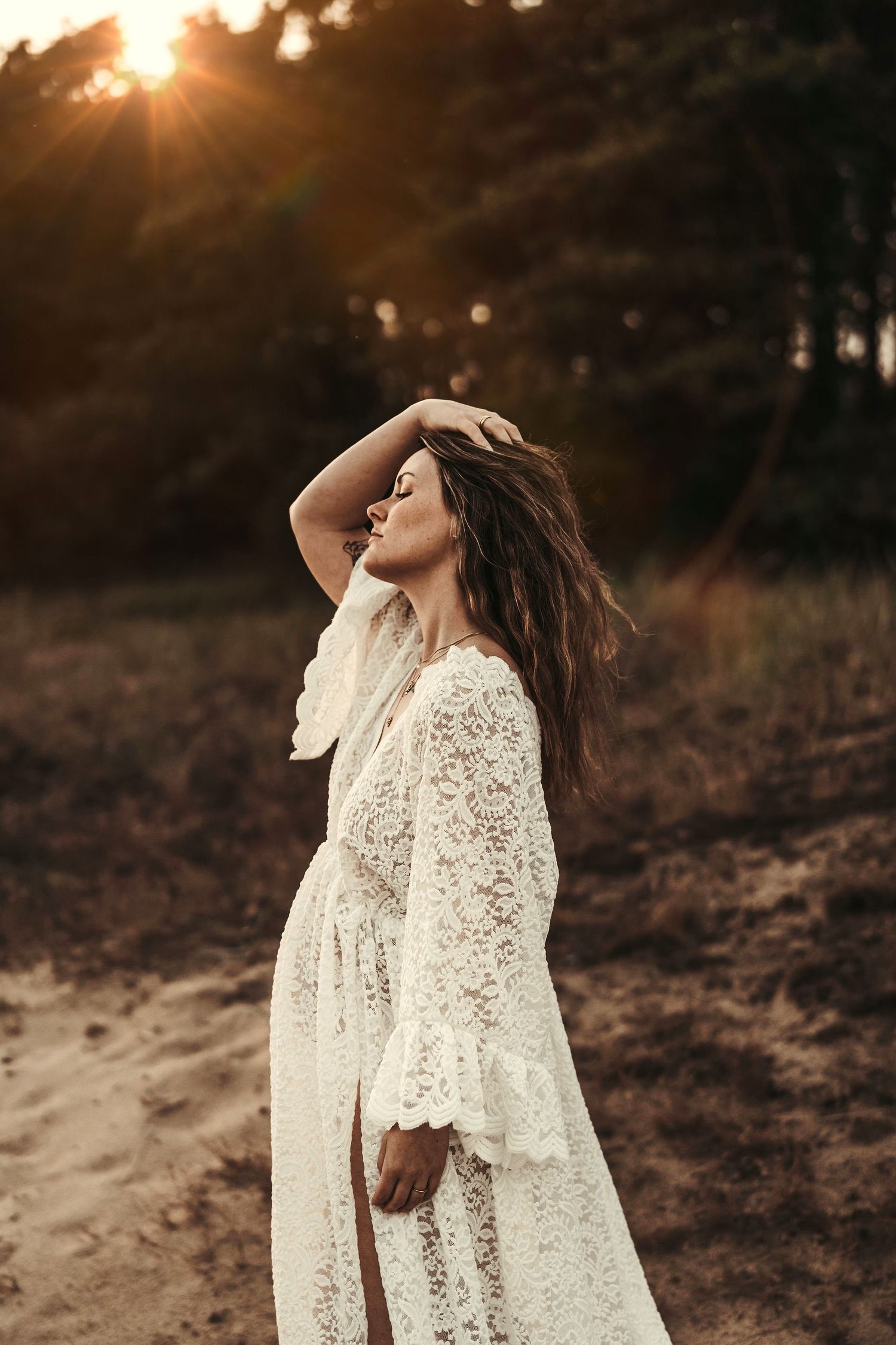 Someone in a delicate white lace dress poses outdoors during golden hour sunset in a forest setting.