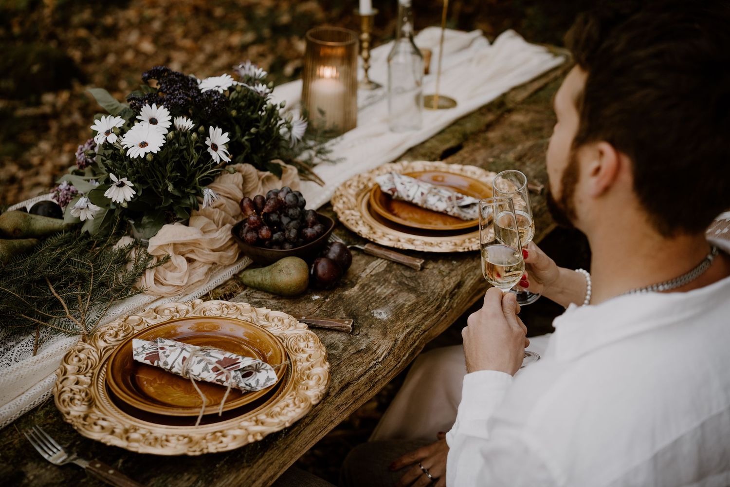 Rustic wedding table setting with gold chargers, linen napkins, and dark floral centerpiece at an outdoor celebration.
