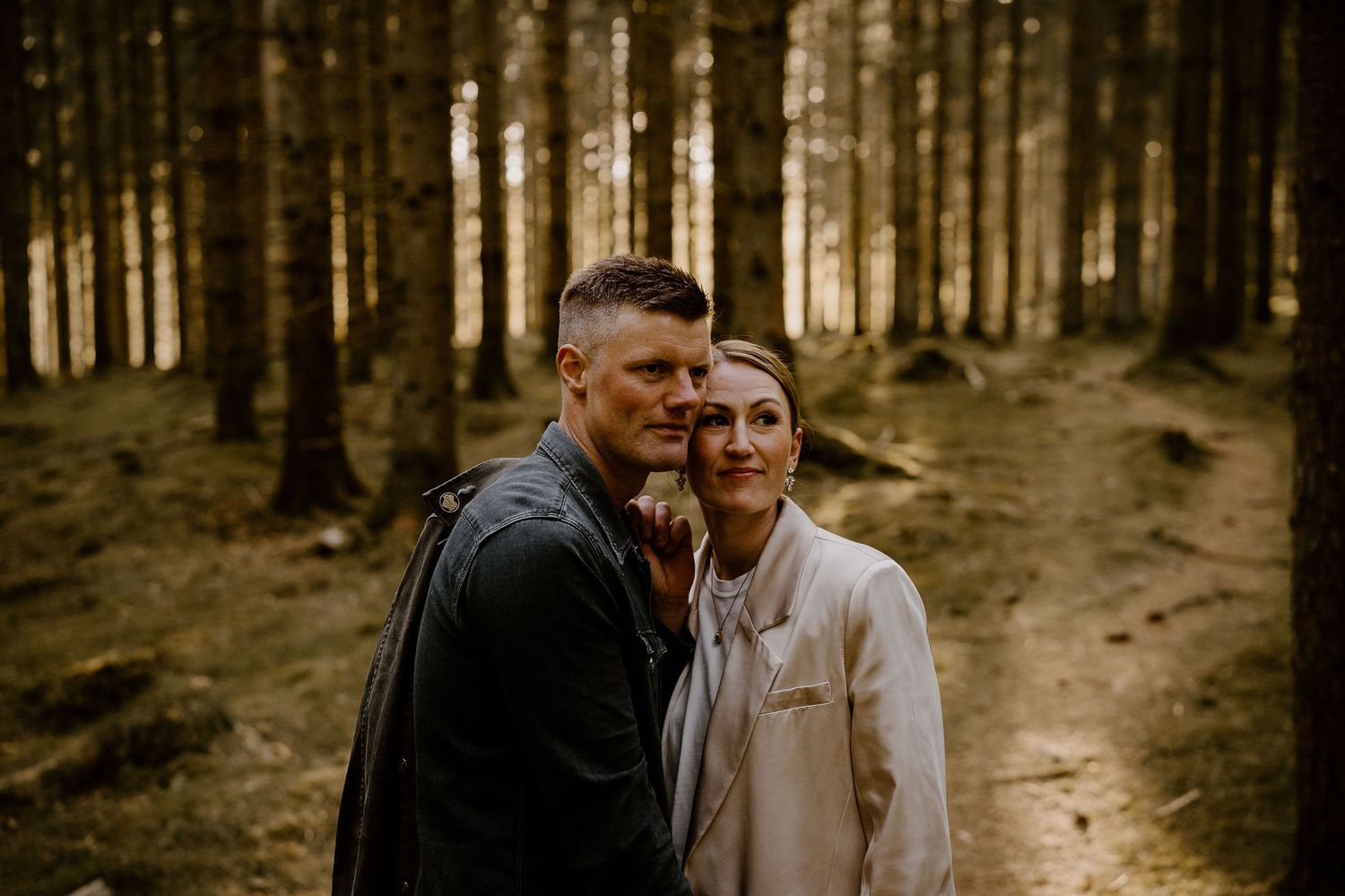 A couple embraces while standing in a moody forest with tall pine trees and golden sunlight filtering through.