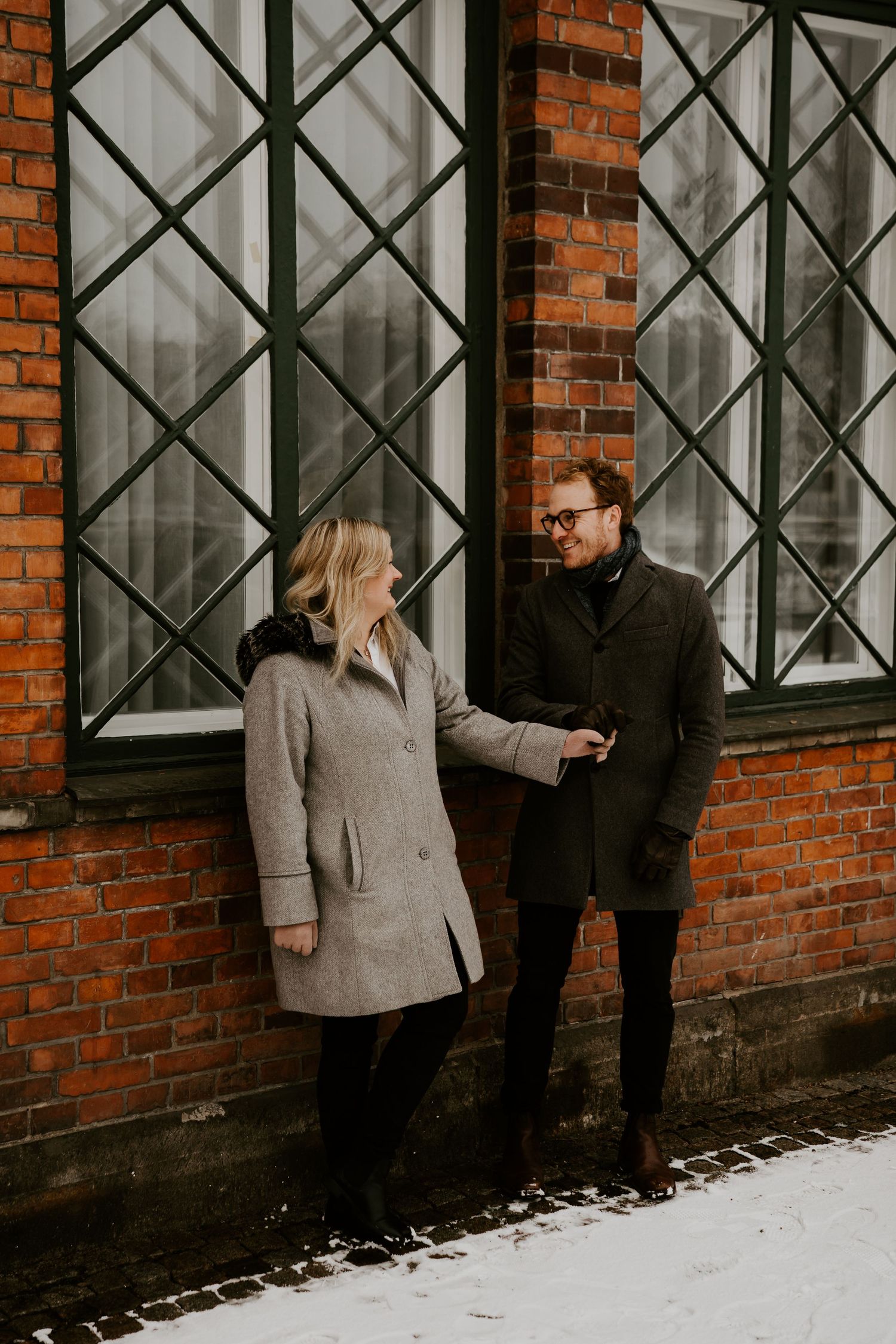 Two people in winter coats having a conversation by a brick building with diamond-paned windows in snowy weather.