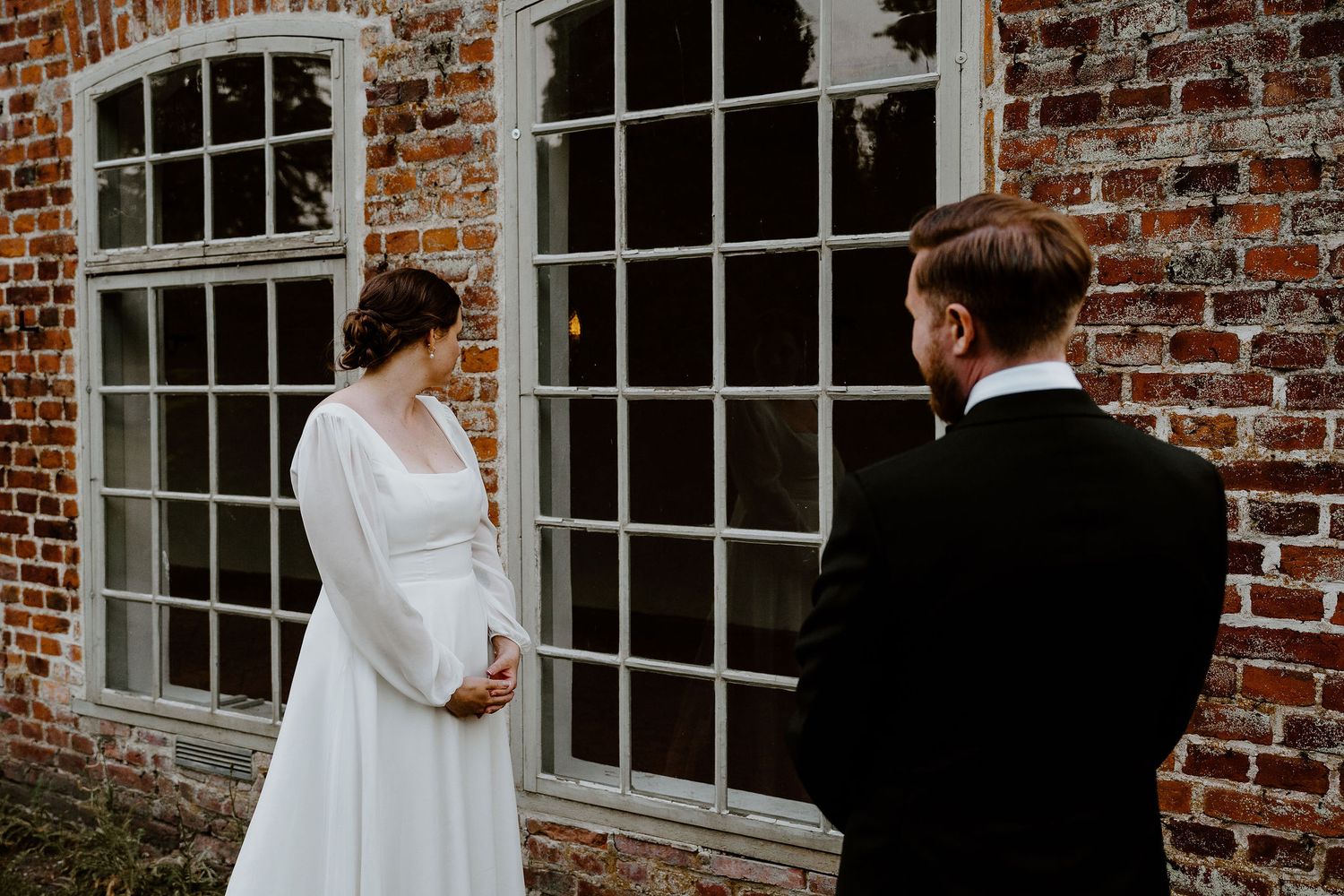 A bride and groom share a moment together outside a rustic brick building with colonial-style windows.