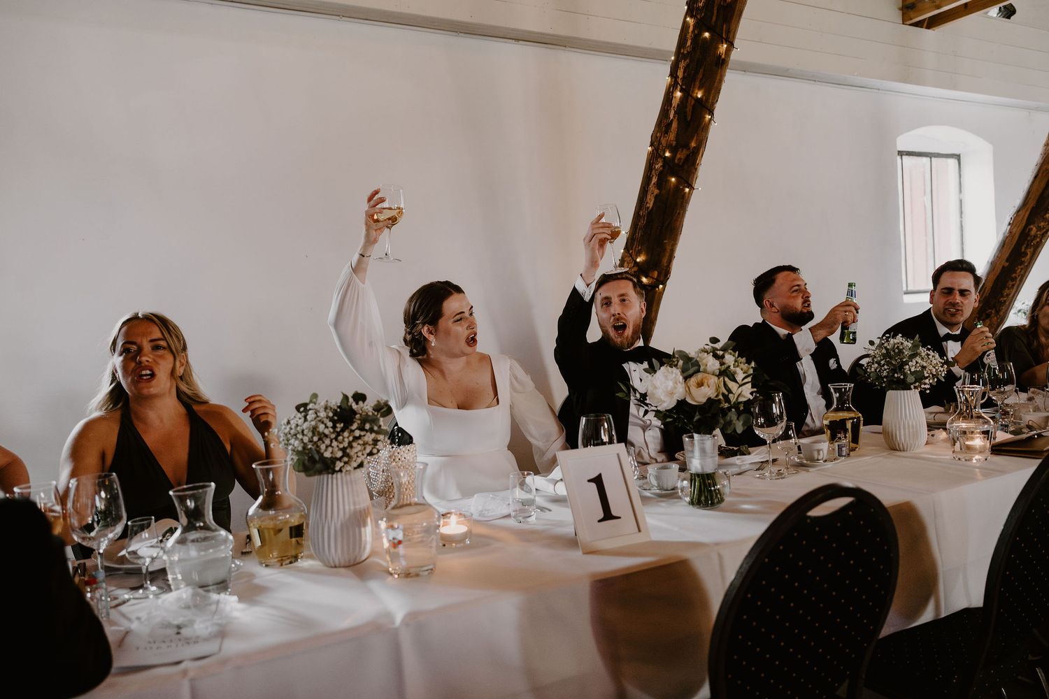 Tables of wedding guests enjoying their dinner in an elegant white reception room with soft lighting and celebratory ambiance.