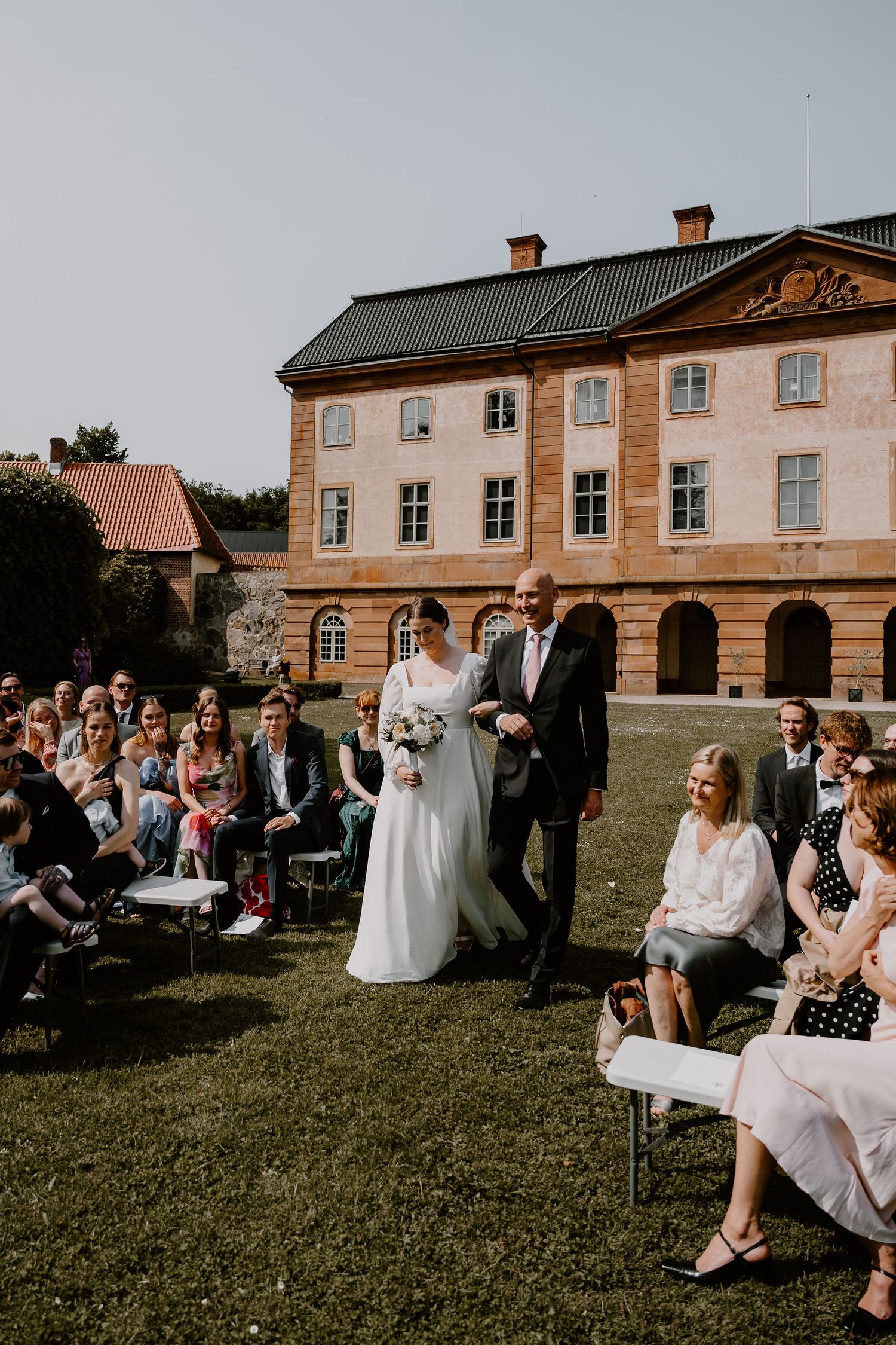 A traditional European manor house with pink-tinted walls hosts an outdoor wedding ceremony on a sunny day.