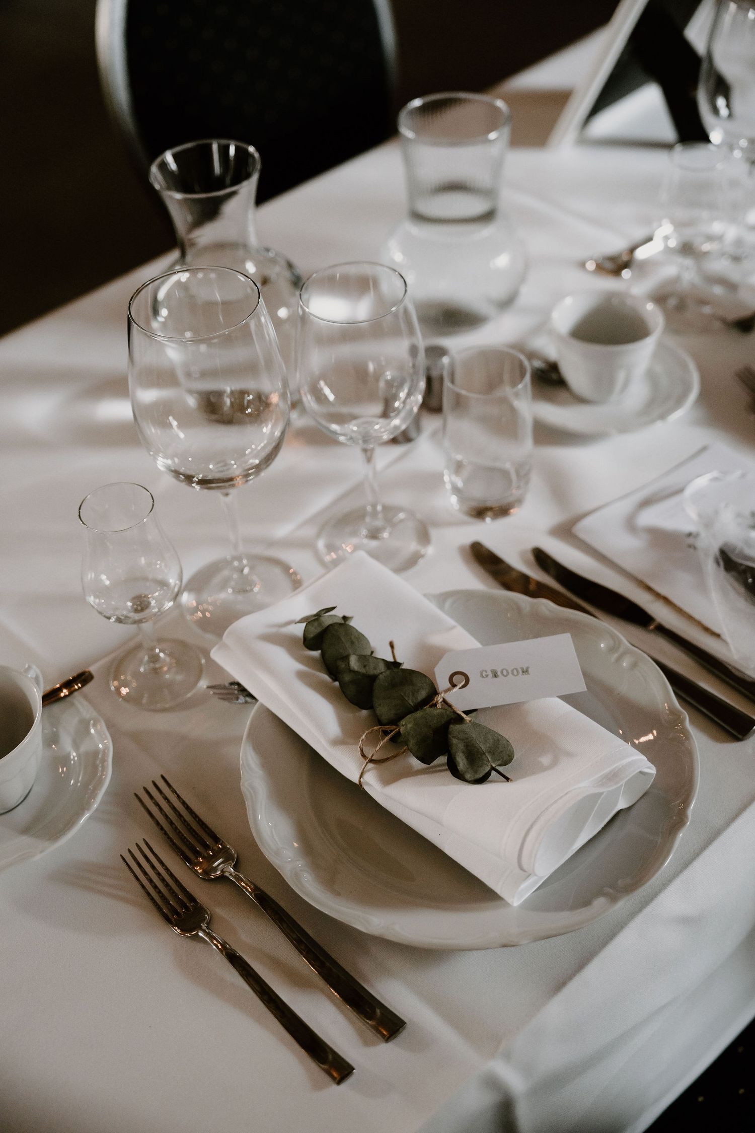 Elegant place setting with wine glasses, white linens, and eucalyptus sprig on formal dining table at upscale restaurant.