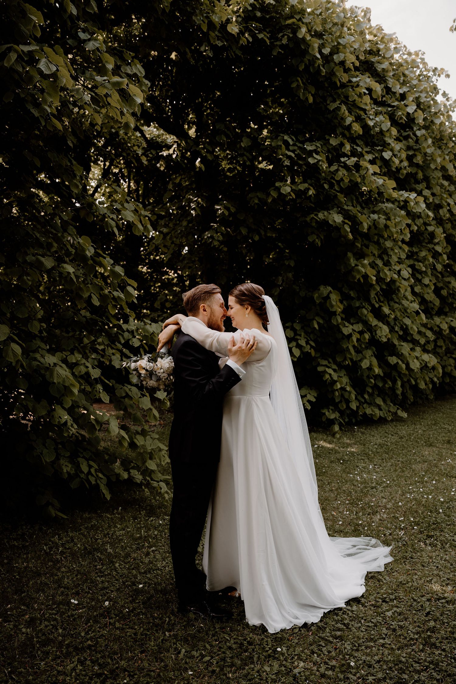 A newlywed couple embraces in an intimate outdoor setting with lush greenery and trees as their backdrop.