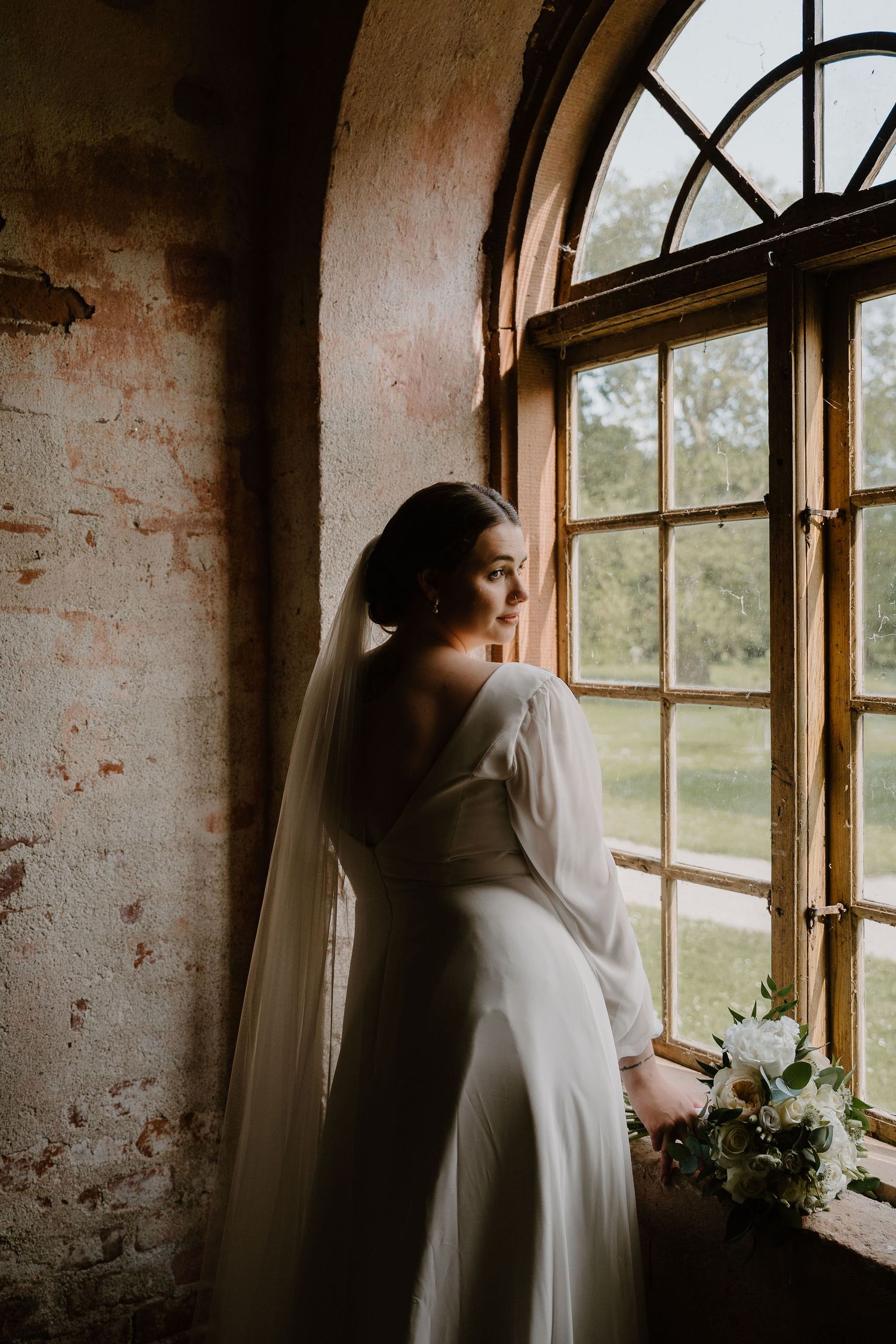 A person in a flowing white dress gazes contemplatively through arched windows in a rustic, sun-lit brick room.