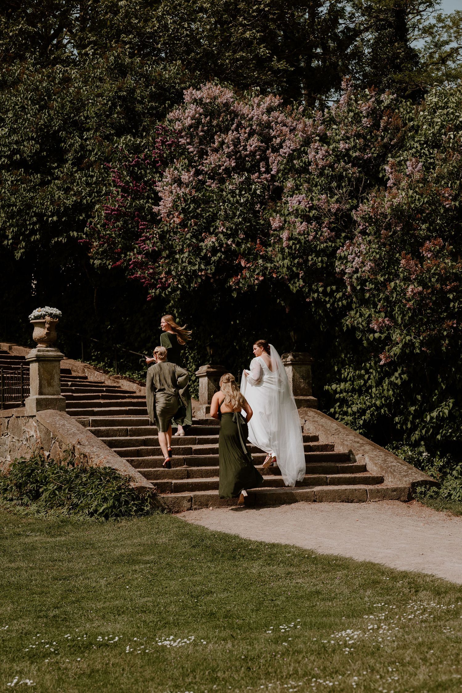 A group walks down elegant stone garden steps surrounded by blooming trees and lush greenery in a romantic outdoor setting.
