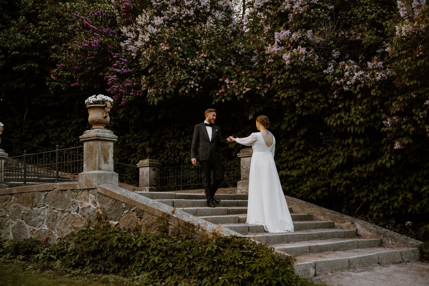 Couple in formal wedding attire descends elegant stone garden steps adorned with decorative urn and blooming foliage.